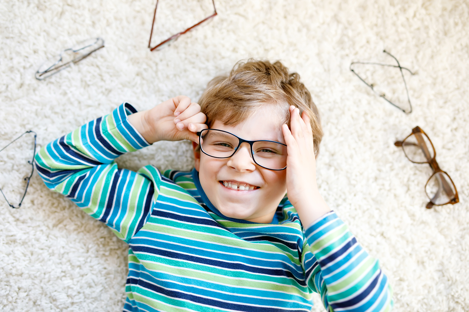 Close-up portrait of little blond kid boy with different eyeglasses on white background. Happy smiling child in casual clothes. Childhood, vision, eyewear, optician store. Boy choosing new glasses
