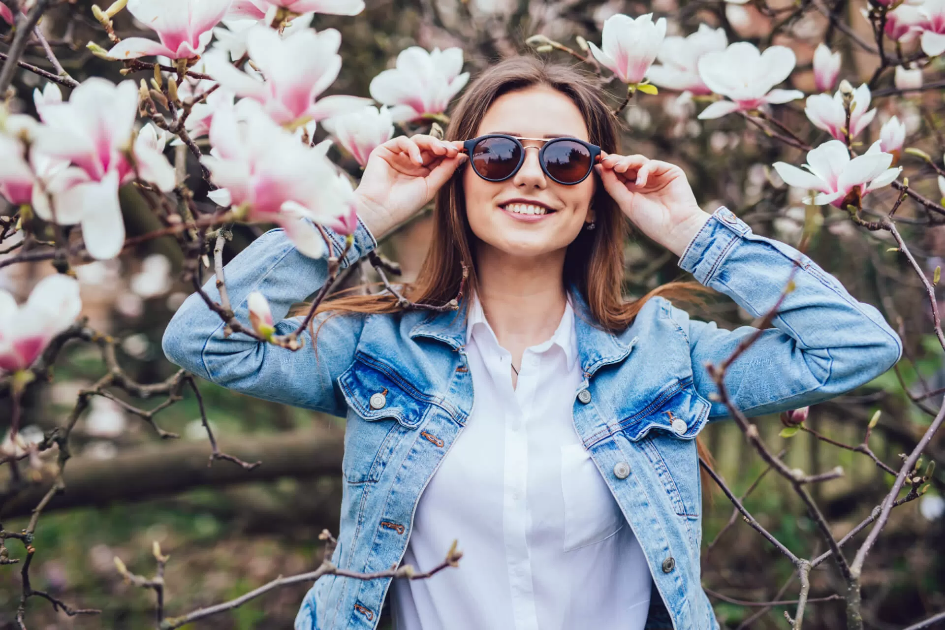 A woman poses between pink flowers while wearing sunglasses.