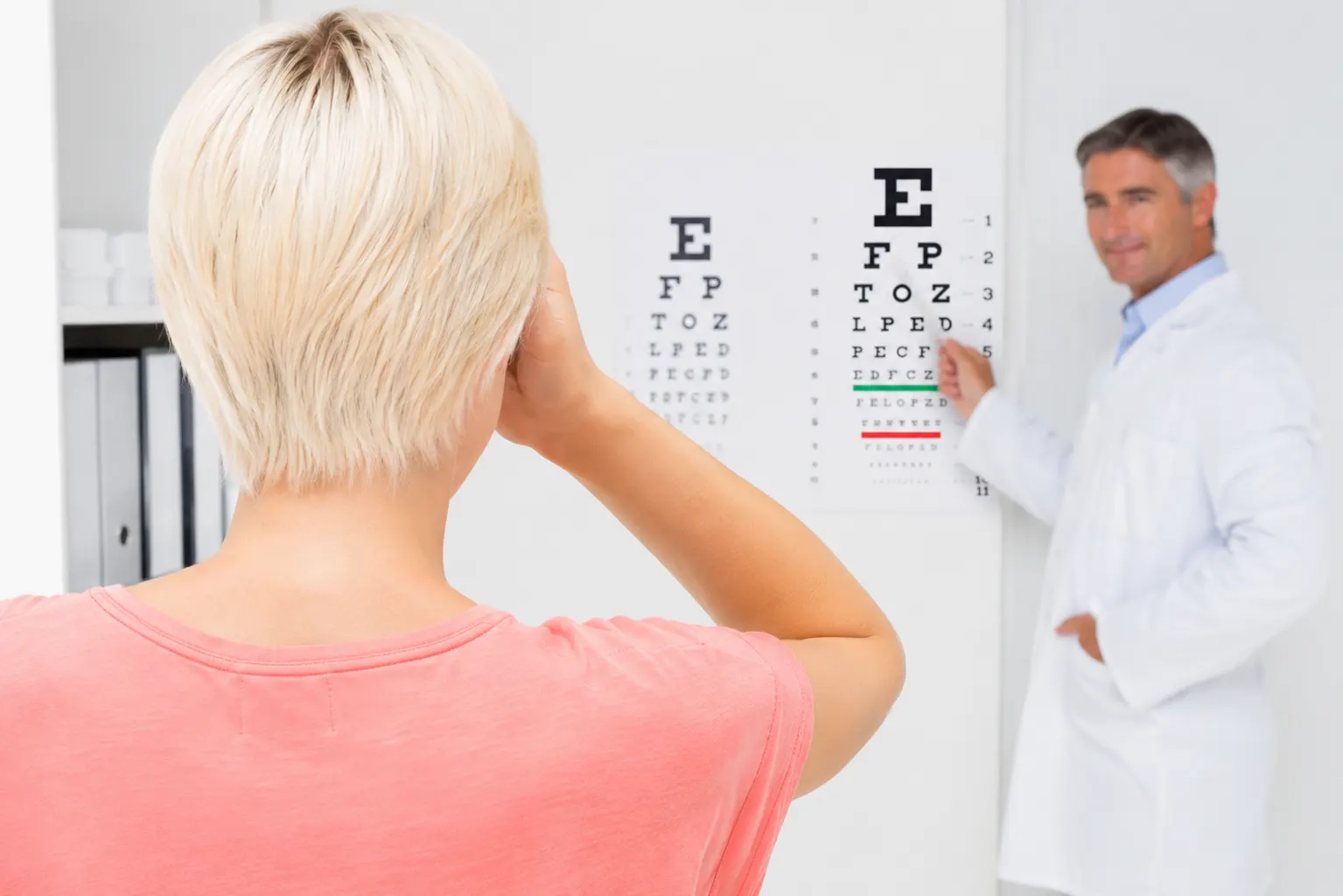 The back of a woman's head can be seen with her covering her right eye with her hand, while an optometrist points to letters for her to read.