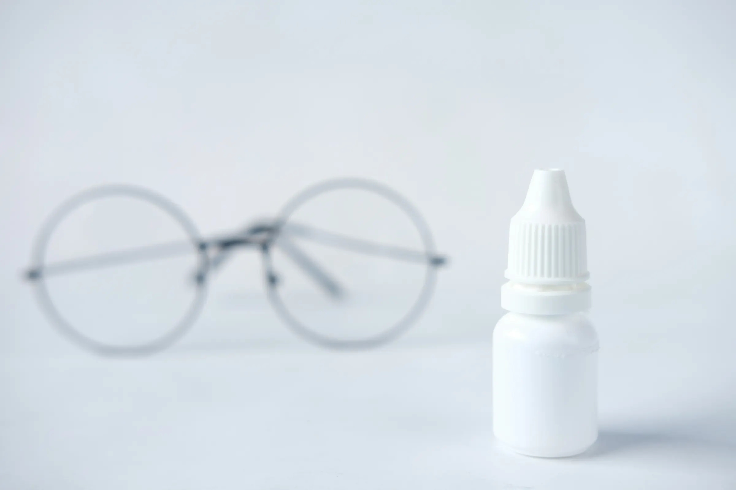 A bottle of eye drops sits in focus on a white table with thin-wired glasses in the background.